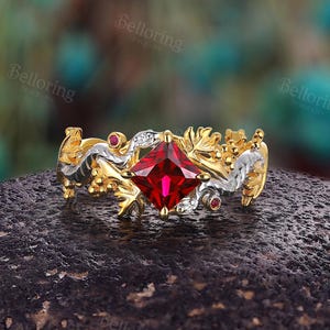 May include: A gold and silver ring with a red gemstone set in a square shape. The ring features intricate floral and leaf designs.