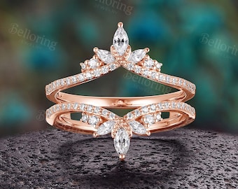 May include: A rose gold ring with a crown-like design, featuring marquise-cut and round-cut clear gemstones. The ring has two bands, with the top band curving upwards to form the crown. The ring is set on a dark, textured surface.