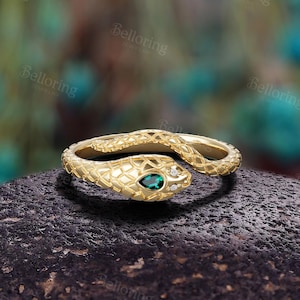 May include: A gold-coloured snake ring with a textured pattern. The snake's head features a pear-shaped green gemstone and a small white gemstone. The ring is set on a dark, textured surface.