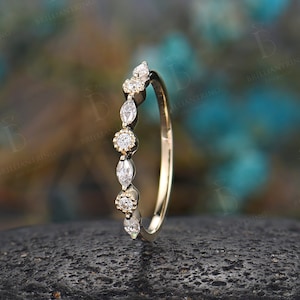 May include: A delicate gold band ring with a row of marquise and round diamonds. The ring is set against a dark grey background.