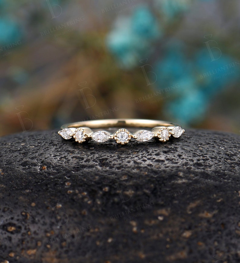 May include: A delicate gold band featuring a row of marquise and round diamonds. The band is set against a dark grey background.