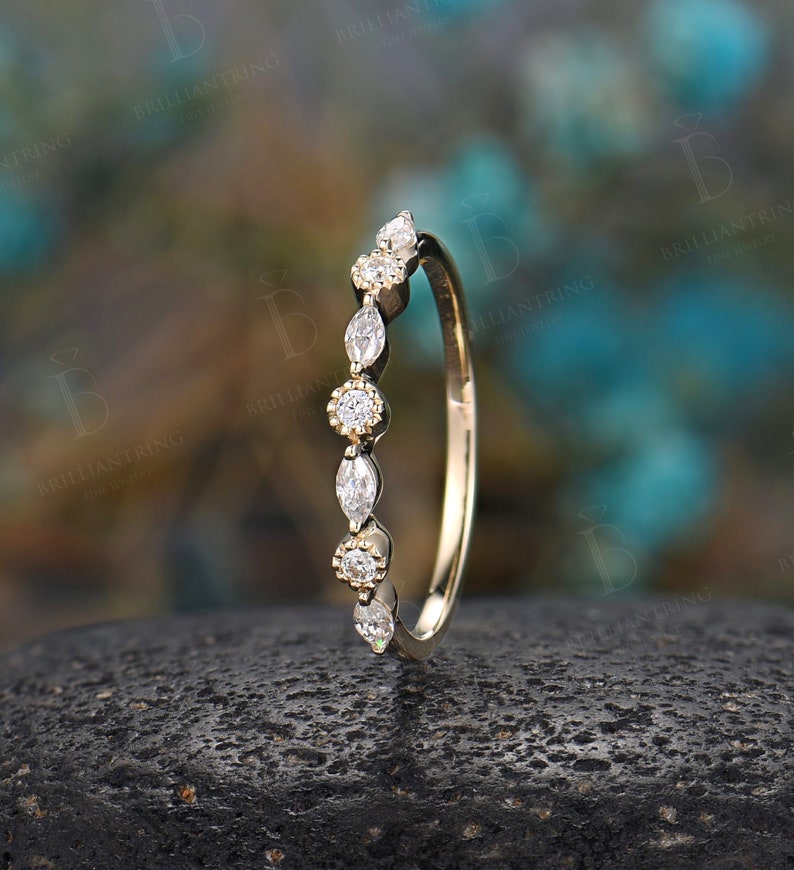 May include: A delicate gold band ring with a row of marquise and round diamonds. The ring is set against a dark grey background.