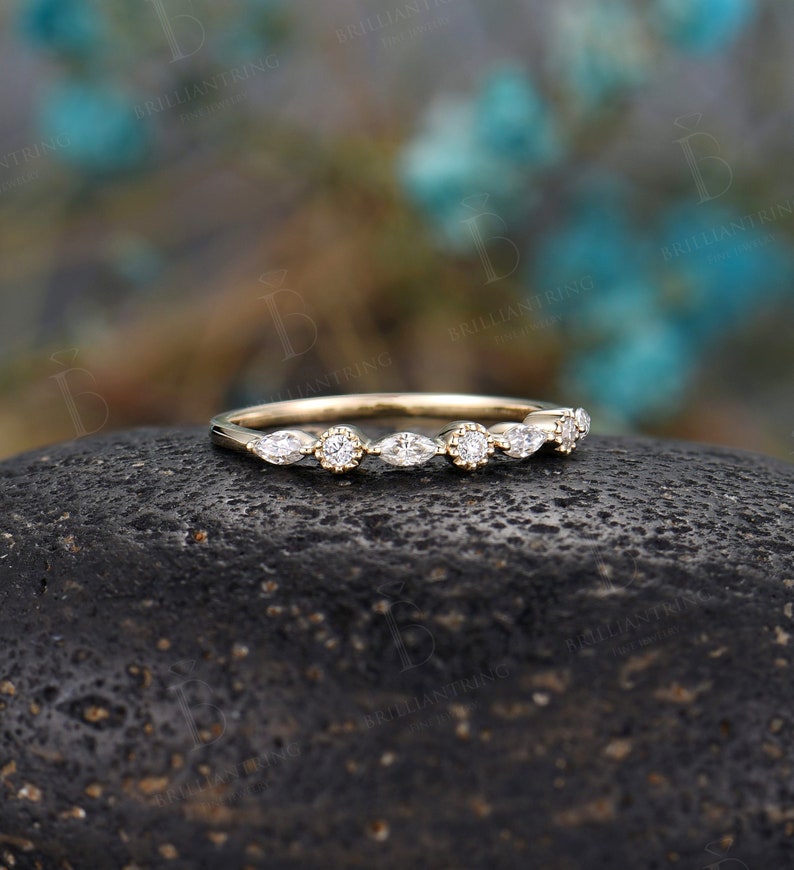 May include: A delicate gold band featuring a row of marquise and round diamonds. The band is set against a dark grey stone.