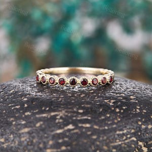 May include: A gold band ring with a series of small, round, deep red gemstones set around the band. The ring is displayed on a dark, textured surface with a blurred green and blue background.