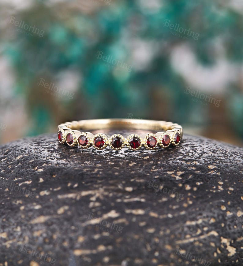 May include: A gold band ring with a series of small, round, deep red gemstones set around the band. The ring is displayed on a dark, textured surface with a blurred green and blue background.