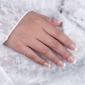 May include: A delicate gold band ring with a row of small, round, clear gemstones. The ring is on a hand with white nail polish.