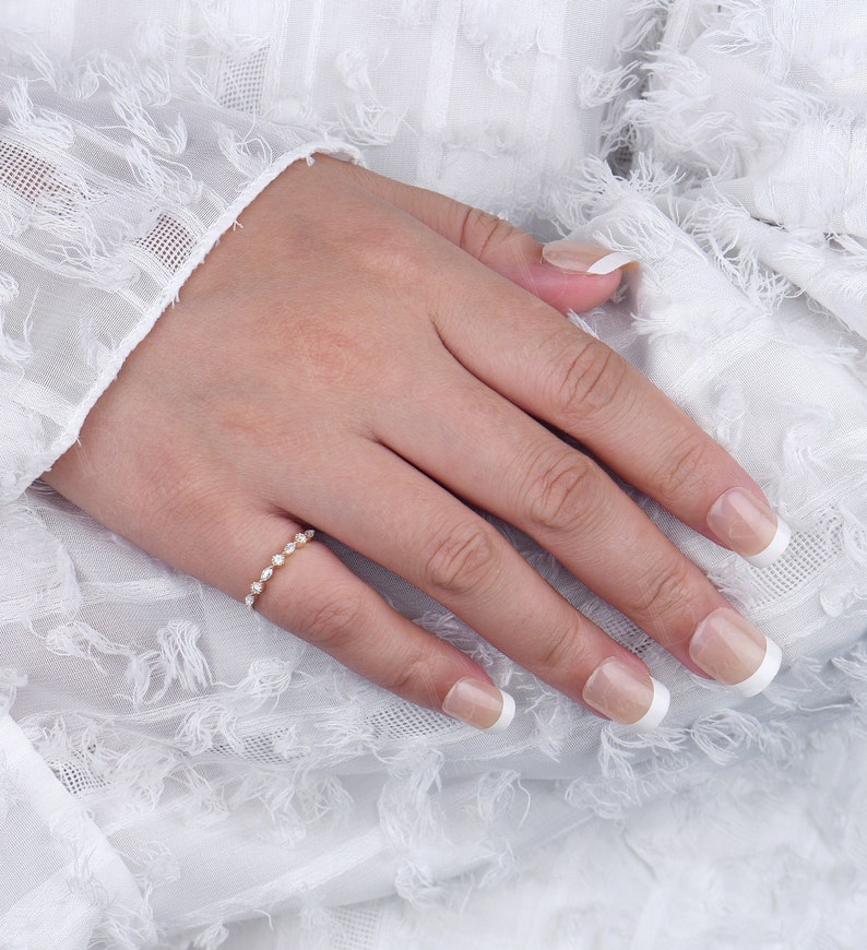 May include: A delicate gold band ring with a row of small, round, clear gemstones. The ring is on a hand with white nail polish.