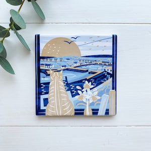 May include: A blue and white ceramic coaster featuring a colourful illustration of a seaside town, a cat, and flowers. The sun is setting over the town.