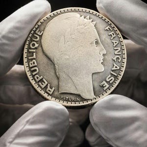 May include: A silver-toned French coin featuring a profile of a figure wearing a laurel wreath. The text REPUBLIQUE FRANÇAISE is embossed along the edge, with the name P. TURIN inscribed at the base of the neck. The coin has a serrated edge.