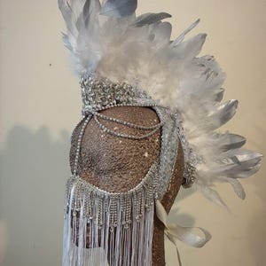 May include: A silver and white feathered headdress with rhinestone and beaded accents. The headdress features a central crest of white feathers, a beaded forehead band, and a draped fringe of white strands.