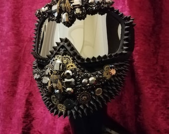 May include: A black and silver steampunk-style mask with a mirrored visor, studded with spikes and embellished with gears and bee charms.
