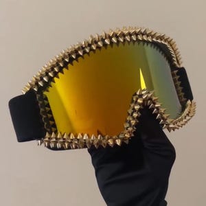 May include: Ski goggles with a gold-colored, spiked frame and a mirrored lens that reflects yellow and green hues. The goggles have a black strap and frame. The image shows a close-up of the goggles.