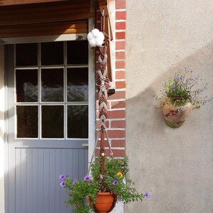 May include: A brown macrame plant hanger with a terracotta pot and purple flowers hanging from a brick wall next to a gray door.