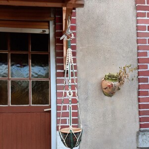 Op de afbeelding: Een macramé plantenhanger met een groen-wit koord, hangend aan een haak aan een bakstenen muur. De hanger houdt een terracotta pot met een groen-wit ontwerp.