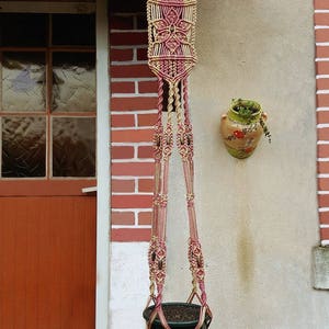 Könnte beinhalten: Ein Makramee-Pflanzenhänger mit einem rosa und beigefarbenen Muster hängt an einer Backsteinwand. Der Hänger hat oben ein Blumendesign und unten einen Topf mit einer Pflanze.
