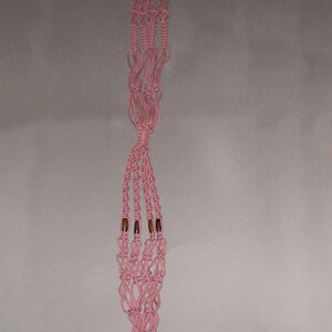May include: A pink macrame plant hanger with wooden beads. The hanger is made of knotted string and has a tassel at the bottom.