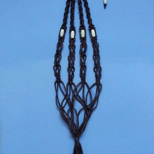 Peut inclure: Un porte-plante en macramé noir avec des perles blanches. Le porte-plante a un pompon en bas.