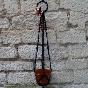 Puede incluir: Un macetero de macramé negro con cuentas de madera y una maceta de terracota. El macetero tiene un diseño de mariposa en la parte superior.