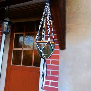 Peut inclure: Un porte-plante en macramé bleu et blanc avec un pot en céramique vert et marron suspendu. Le porte-plante est fixé à une poutre en bois au-dessus d'une porte rouge.