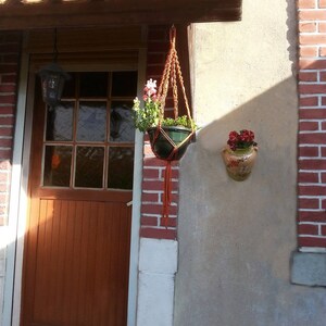 Peut inclure: Un porte-plante en macramé suspendu avec un pot en terre cuite rempli de fleurs roses. Le porte-plante est fait de corde en fibre naturelle et est suspendu à une poutre en bois. Le pot est suspendu devant un mur en brique et une porte en bois.
