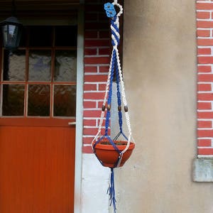 Könnte beinhalten: Ein blauer und weißer Makramee-Pflanzenhänger mit einem Terrakotta-Topf, der daran hängt. Der Hänger ist an einer Backsteinwand mit weiß gestrichener Oberfläche befestigt.