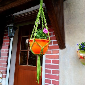 May include: A green macrame plant hanger with a bright orange pot and pink flowers hanging from a brick wall.