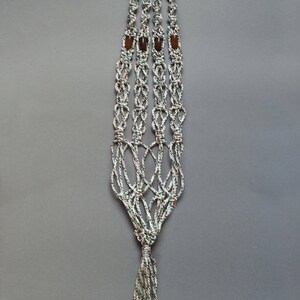 Peut inclure: Un porte-plante en macramé blanc et vert avec des perles en bois brun. Le porte-plante a un long pompon en bas.