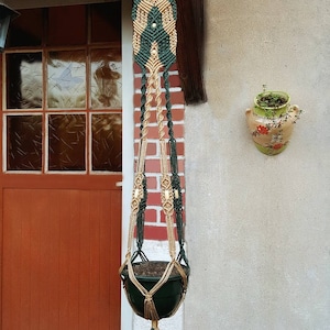 Peut inclure: Un porte-plante en macramé vert et beige avec un petit pot vert suspendu. Le porte-plante est fixé à un mur en briques et possède un pompon en bas.