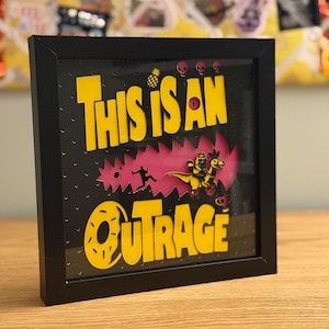 May include: A framed art piece with the words "THIS IS AN OUTRAGE" in yellow, set against a black background. The design includes a pink burst, a running figure, and a dinosaur riding a vehicle. Small skulls and a pineapple are also featured.