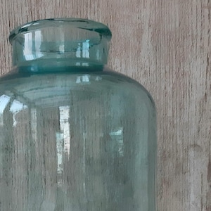 May include: A large, clear glass jar with a wide, cylindrical body and a slightly narrower neck. The jar has a teal-blue tint and sits on a white surface against a weathered wood background. The jar is empty.