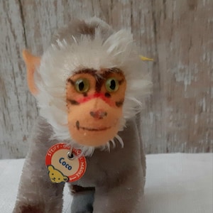 May include: A vintage Steiff Coco monkey plush toy. The plush monkey has a brown body, white fur around the face, and a red and orange face. It has a red tag that says "Coco".