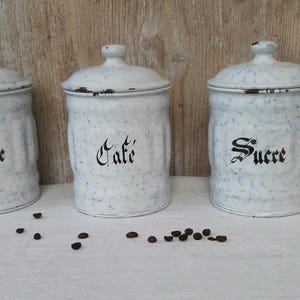 Könnte beinhalten: Drei weiße Emaille-Vorratsdosen mit blauem Sprenkelmuster und passenden Deckeln. Jede Dose ist mit schwarzer, dekorativer Schreibschrift beschriftet: Chicorée, Café und Sucre. Vor den Dosen liegen einige dunkle Kaffeebohnen verstreut.