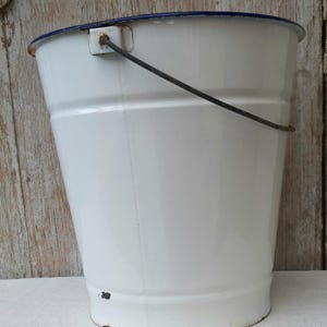 May include: A white enamel bucket with a blue rim and a metal handle. The bucket has a slightly tapered shape and shows signs of wear, with some rust visible on the bottom. The handle is attached with metal brackets.
