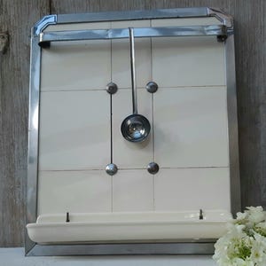 May include: A rectangular wall-mounted kitchen rack featuring a chrome metal frame, a white tiled back panel with four decorative metal studs, a horizontal chrome bar at the top, a matching white shelf at the base, and a hanging metal ladle.