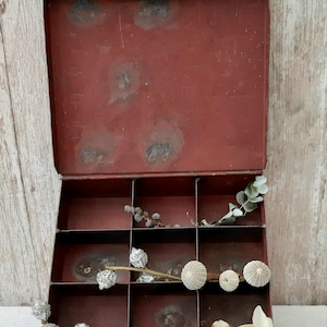 Könnte beinhalten: Eine offene, antike rote Metallbox mit neun Fächern. Die Box ist mit getrockneten Pflanzen und weißen Blumen dekoriert. Sie hat einen Klappdeckel und einen kleinen Riegel. Die Box steht auf einer weißen Oberfläche.