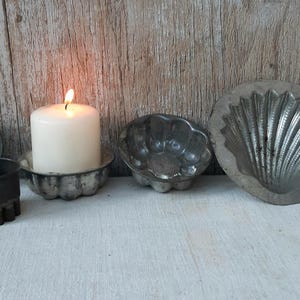 Op de afbeelding: Een verzameling vintage metalen mallen en een kaars op een wit oppervlak. De mallen hebben verschillende vormen, waaronder een rechthoekige, een ronde en een schelpvormige. Een brandende witte kaars staat in een decoratieve houder, met gedroogde bloemen rechts.