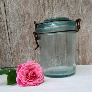 Op de afbeelding: Een doorzichtige, lichtblauwe glazen voorraadpot met een bijpassend glazen deksel, vastgezet met een roestig metalen beugelsluiting. Een enkele, felroze roos met groene bladeren is direct voor de glazen container geplaatst.