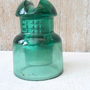 May include: A vintage, emerald green glass insulator. The insulator has a rounded base, a cylindrical body, and a top with a groove. The glass has a slight texture and a glossy finish. The insulator is likely from the early 20th century.