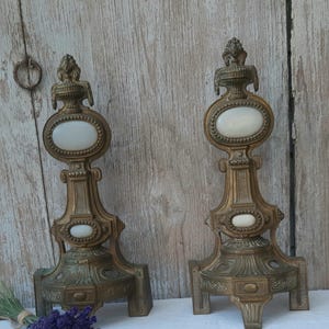 May include: A pair of matching brass fireplace andirons featuring ornate metalwork, a wide base with three feet, and two oval-shaped white glass inlays on each piece. The vertical structures are topped with decorative finials and intricate detailing.