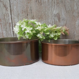Peut inclure: Deux casseroles anciennes en cuivre avec de longues poignées en métal foncé. L'une a une teinte brun-verdâtre ternie, l'autre est en cuivre brillant. Un petit bouquet de fleurs blanches et roses est placé entre elles.