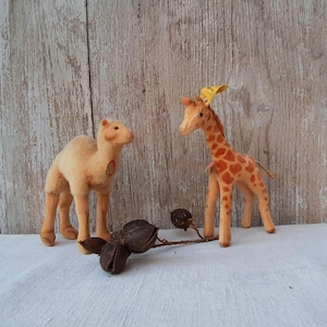 Op de afbeelding: Een beige kameel en een oranje giraf met bruine vlekken staan op een wit oppervlak. Een takje met gedroogde zaaddozen ligt op de voorgrond. De achtergrond is licht hout. Handgemaakte speelgoed.