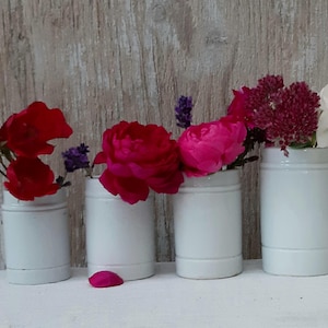 Könnte beinhalten: Vier weiße Keramikvasen, jede mit einem anderen Blumenarrangement. Die Blumen umfassen rote, rosa, lila und weiße Blüten. Die Vasen sind zylindrisch und stehen auf einer weißen Oberfläche vor einem Holzgrund.