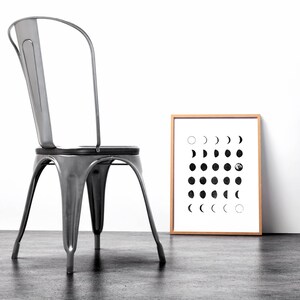 May include: A black and white print of the moon phases in a wooden frame, hanging on a white wall next to a silver metal chair.