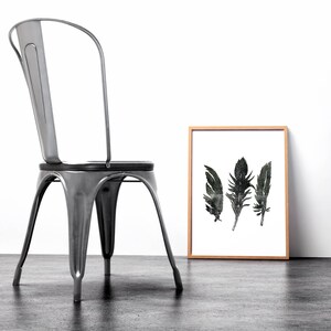 May include: A black and white print of three feathers in a wooden frame, sitting on a grey floor next to a silver metal chair.