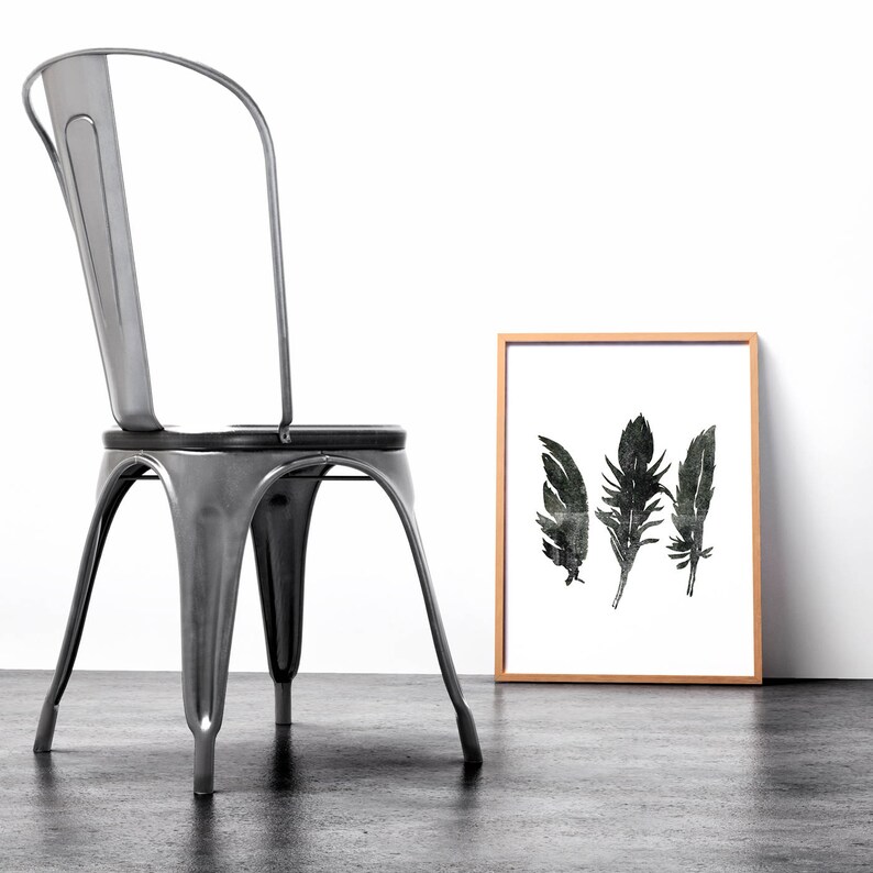 May include: A black and white print of three feathers in a wooden frame, sitting on a grey floor next to a silver metal chair.
