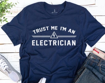 May include: A navy blue t-shirt with the text "Trust me I'm an Electrician" in white lettering with a lightning bolt symbol. The shirt is laid flat on a white wooden surface with a pair of grey trainers and blue jeans.