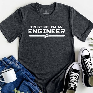 May include: A dark grey t-shirt with the text "Trust me, I'm an Engineer" in white lettering. The shirt has a distressed look and features two small gear icons.