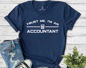 Puede incluir: Una camiseta azul marino con texto blanco que dice "Trust me, I'm an Accountant" con un icono de calculadora. La camiseta está colocada sobre una superficie de madera con un par de zapatos Converse negros y unos vaqueros azules.