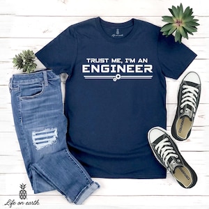 May include: A navy blue t-shirt with white text that reads "Trust me, I'm an Engineer" with two gears underneath the text. The shirt is laid out on a white wooden surface with a pair of blue jeans and black sneakers.