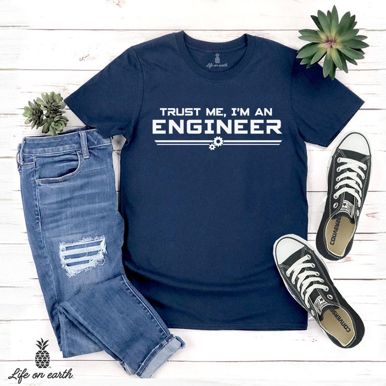 May include: A navy blue t-shirt with white text that reads "Trust me, I'm an Engineer" with two gears underneath the text. The shirt is laid out on a white wooden surface with a pair of blue jeans and black sneakers.